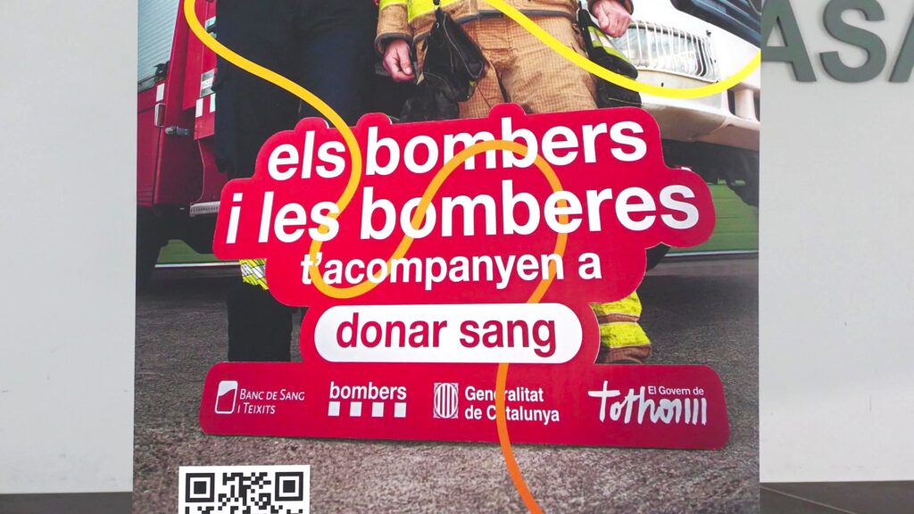 Cartell promocional de la campanya "Els bombers i les bomberes t'acompanyen a donar sang" davant d'un edifici a Amposta, Terres de l'Ebre, amb la presència parcial de dos bombers amb uniforme.