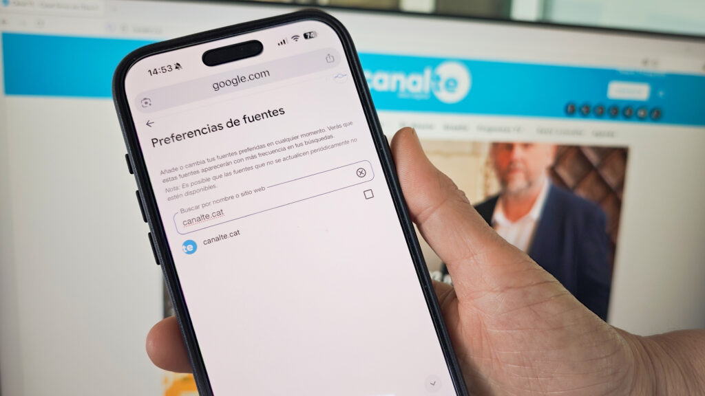 Una mà subjectant un telèfon mòbil que mostra la pantalla de configuració de 'Font Preferida' de Google, amb la web de Canal TE ja seleccionada com a opció, davant d'una pantalla d'ordinador de fons a Tortosa, Terres de l'Ebre.