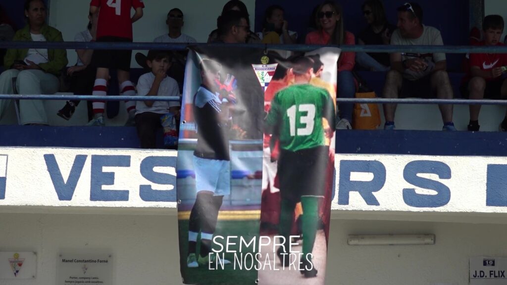 Pancarta d'homenatge a Manel Constantino Forné penjada a la grada del camp d'esports de Masdenverge amb públic a les Terres de l'Ebre