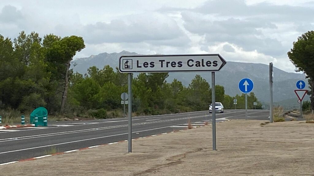 Senyal de trànsit indicant l’entrada a la urbanització de les Tres Cales, a l’Ametlla de Mar, situada al marge d’una carretera envoltada de vegetació, amb muntanyes i un cel ennuvolat al fons.