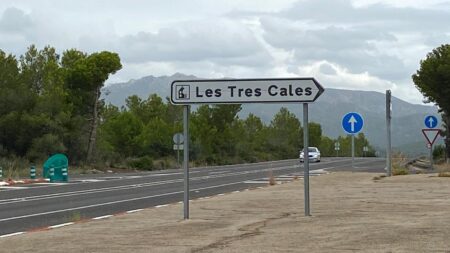 Senyal de trànsit indicant l’entrada a la urbanització de les Tres Cales, a l’Ametlla de Mar, situada al marge d’una carretera envoltada de vegetació, amb muntanyes i un cel ennuvolat al fons.